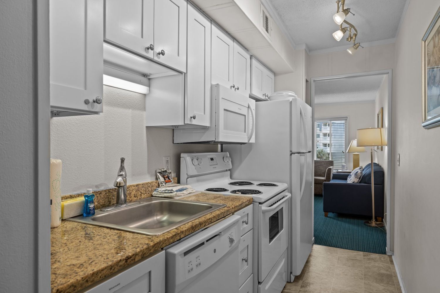 A compact kitchen with white cabinets, a stove, and a refrigerator. There is a sink with a faucet, and a countertop with a dish soap bottle. A dining area with a blue carpet and a lamp is visible in the background.