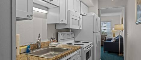 A compact kitchen with white cabinets, a stove, and a refrigerator. There is a sink with a faucet, and a countertop with a dish soap bottle. A dining area with a blue carpet and a lamp is visible in the background.