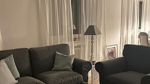 A living room with a dark brown sofa, a side table, a floor lamp, and a window with sheer curtains. - Feel-good vacation in Baden-Baden: Your vacation apartment (Baden-Baden)