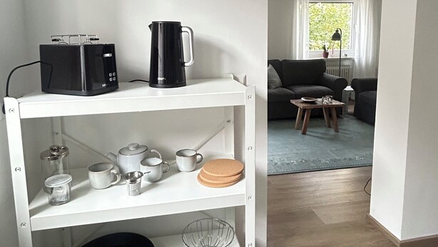 A white shelf with kitchenware, a coffee maker, and a kettle in a room with a wooden floor and a view of a living area with a sofa and a table. - Feel-good vacation in Baden-Baden: Your vacation apartment (Baden-Baden)