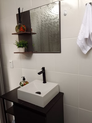 Casa de banho com lavatório branco, torneira preta e espelho com padrão hexagonal. Existem duas prateleiras com objetos decorativos e uma toalha pendurada na parede.