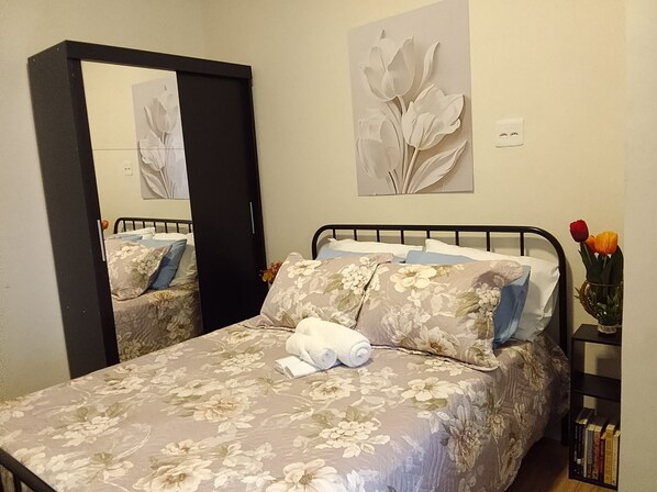 Uma cama bem arrumada com roupa de cama florida, um espelho e um vaso com flores sobre uma mesa de cabeceira.