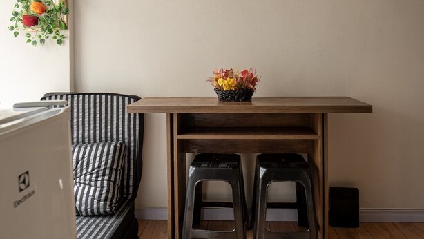 A small, cozy room with a wooden table, two black stools, a striped sofa, and a wall-mounted plant. - Cozy studio in Glória, close to the Lapa arches and Glória metro station. (Rio de Janeiro)