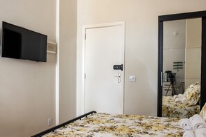 Quarto de hotel com cama, televisão presa na parede, porta e espelho refletindo parte do espaço.