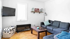 A living room with a grey sofa, a wooden coffee table, a flat-screen TV, a fireplace, and a wall-mounted shelf with decorative items. - Lake Access - Private Spa - Pool table | Rustique (Saint-Claude)