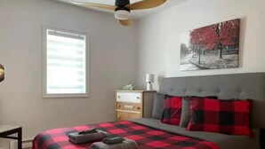 A bedroom with a bed, a nightstand, a dresser, a ceiling fan, a window with blinds, and a wall-mounted painting. - Lake Access - Private Spa - Pool table | Rustique (Saint-Claude)