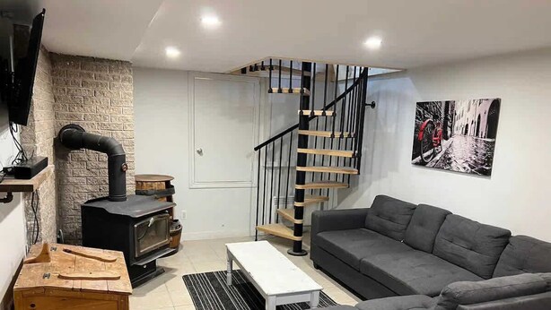 A living room with a gray sofa, a wooden chest, a white coffee table, a black fireplace, and a spiral staircase. - Lake Access - Private Spa - Pool table | Rustique (Saint-Claude)