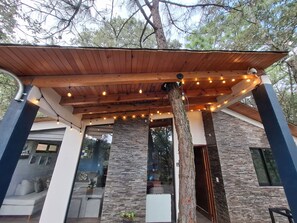 A covered outdoor area with a wooden ceiling and string lights, a stone wall, and a tree trunk.