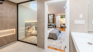 Une chambre moderne avec une cloison en verre, un grand lit, un bureau et une salle de bain dotée d’un lavabo blanc.