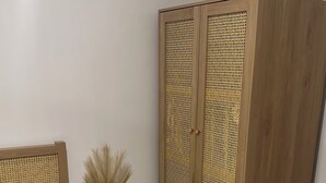 Ein Schlafzimmer mit einem Bett, einem Holzschrank mit Flechtfront, einer Vase mit Trockenpflanzen und zwei Teppichen.