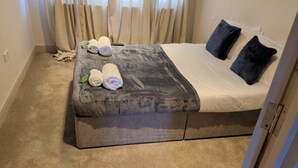 A hotel room with a bed, a bedside table with towels, a dark-colored bench, and a curtain.