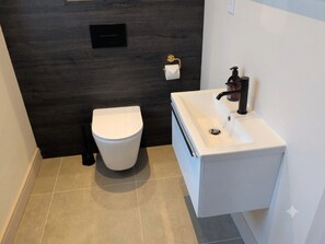 Ein modernes Badezimmer mit Toilette in Weiß, einem Waschbecken mit schwarzem Wasserhahn und einer dunklen Wand.