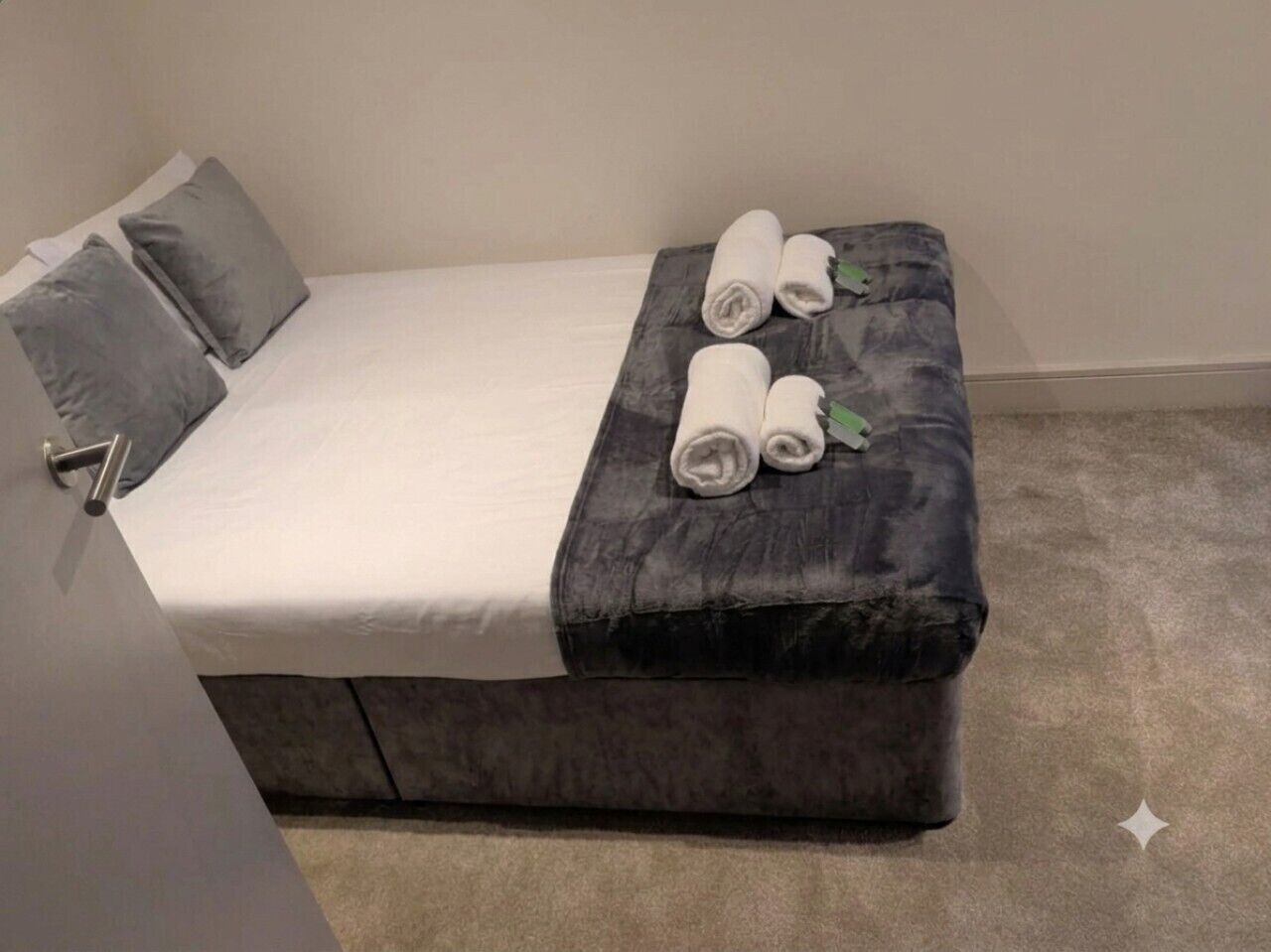 A small, single-bed room with a grey upholstered bench, a white bedspread, and rolled towels on the bench.