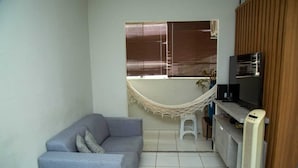Un petit salon avec un canapé gris, un hamac, une télévision et un ventilateur.