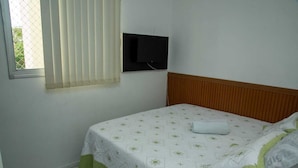 Une chambre à coucher avec un lit, une fenêtre avec des stores, un téléviseur fixé au mur et une tête de lit en bois.