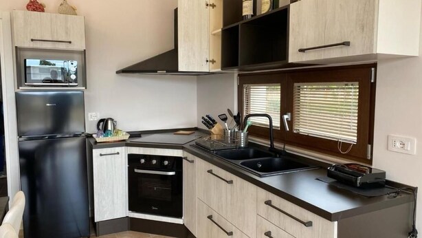 A compact kitchen with a built-in oven, microwave, and a black refrigerator. There are wooden cabinets with black handles, a black countertop, and a window with blinds. - Villa among olive trees with garden and parking • Relax in Puglia (Puglia)