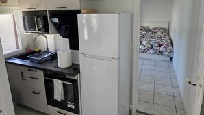 Cocina compacta con microondas, horno y refrigerador. Se ve un lavamanos pequeño con un escurreplatos y un rollo de toallas de papel. En el fondo, una cama con ropa de cama estampada.