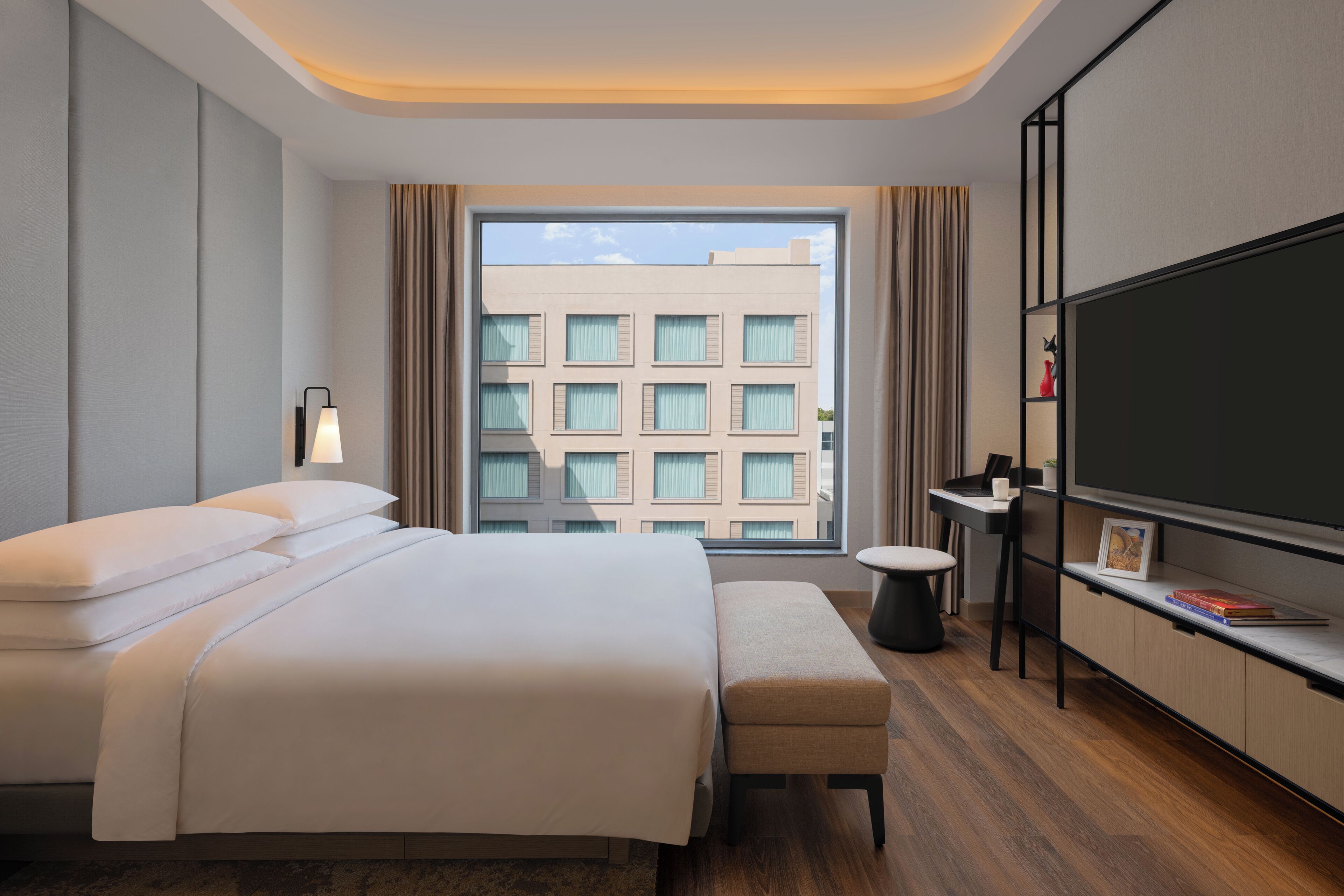 Quarto de hotel moderno com uma cama grande, televisão de tela plana, uma pequena mesa e uma janela com vista para prédios.