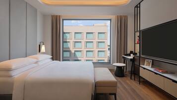 Quarto de hotel moderno com uma cama grande, televisão de tela plana, uma pequena mesa e uma janela com vista para prédios.