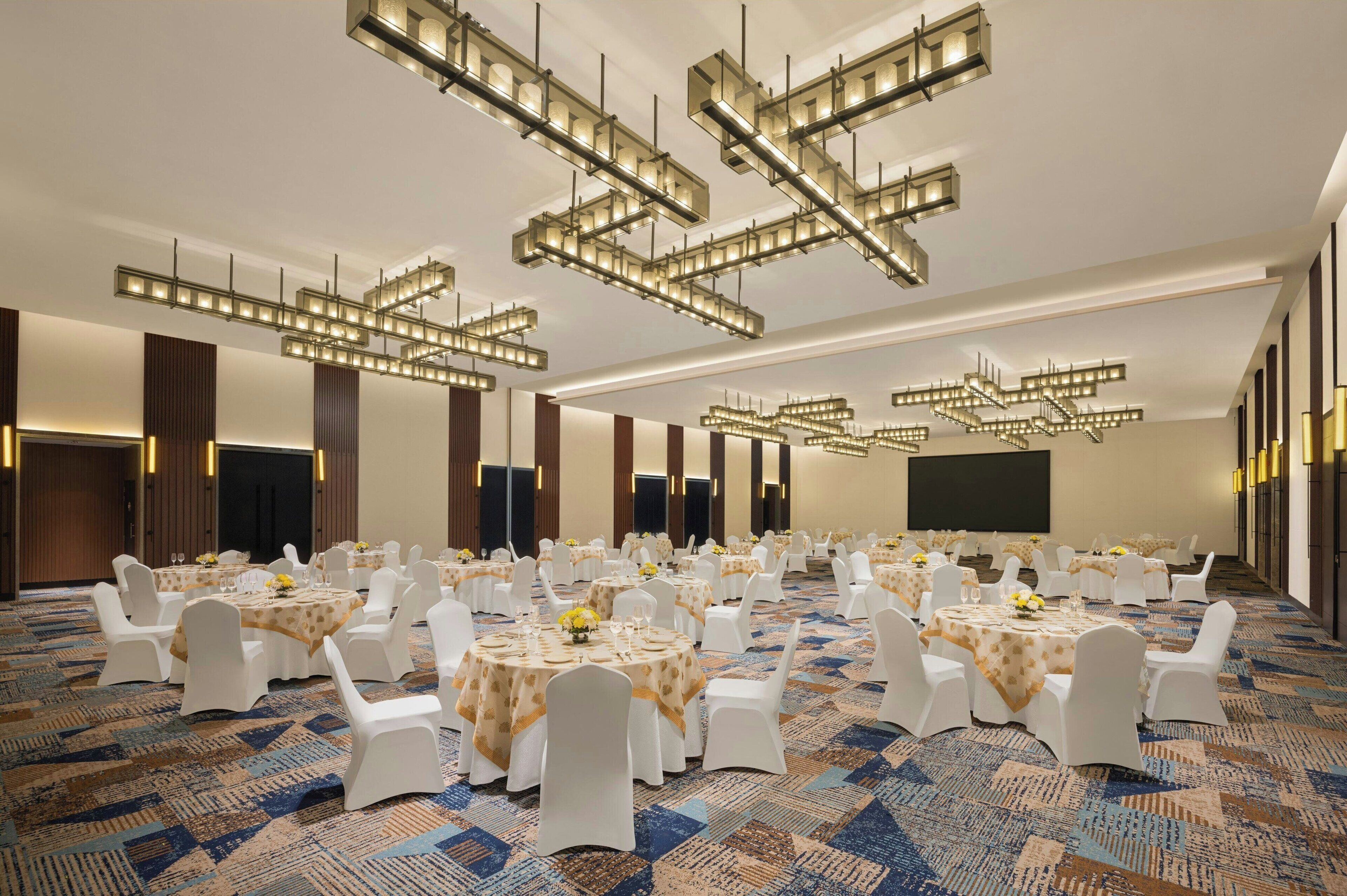 A large banquet hall with round tables, white chairs, and modern chandeliers.