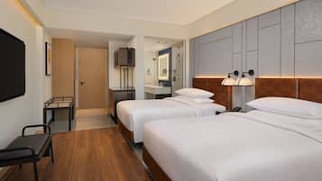 Quarto de hotel com duas camas, uma escrivaninha, uma cadeira, televisão e casa de banho visível através de uma porta aberta.
