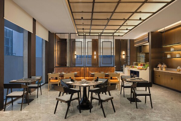 A modern dining area with round tables and chairs, a kitchen area with a counter, and a view through a window. - Four Points By Sheraton Amritsar, Mall Road (Amritsar)