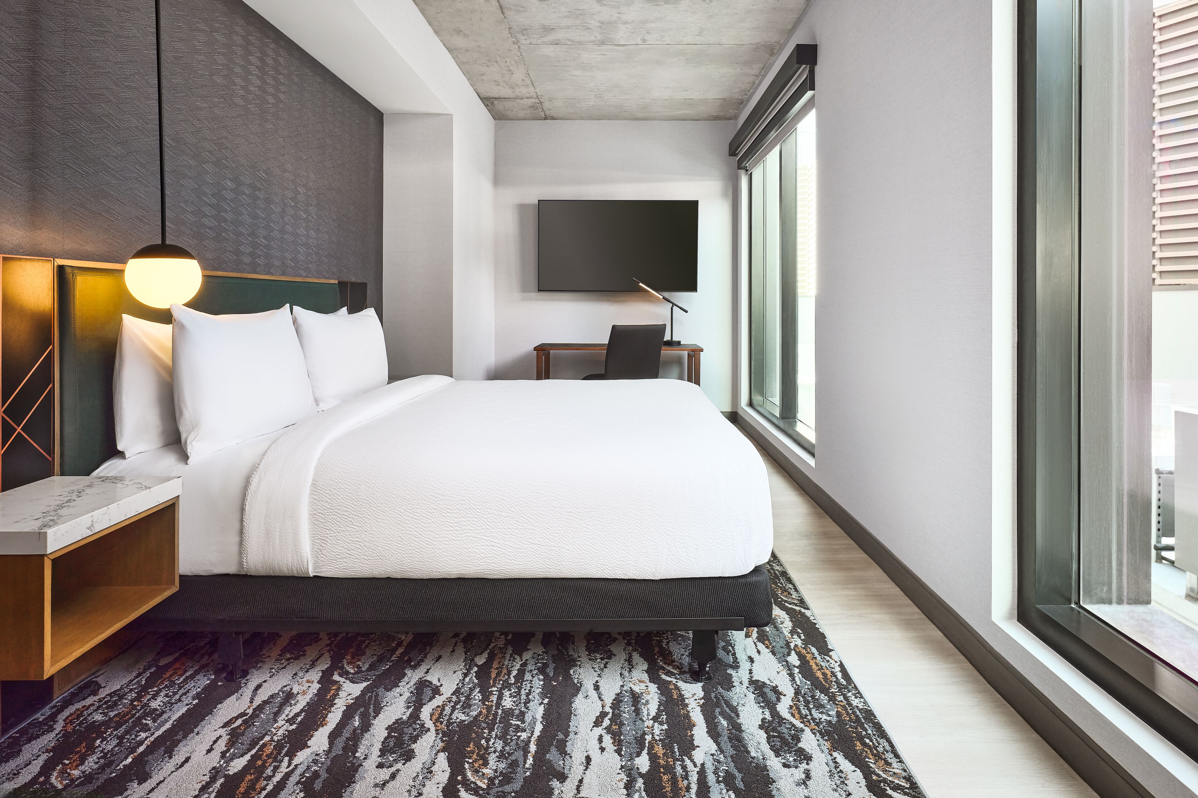 A modern hotel room with a large bed, a desk, a flat-screen TV, and a large window.