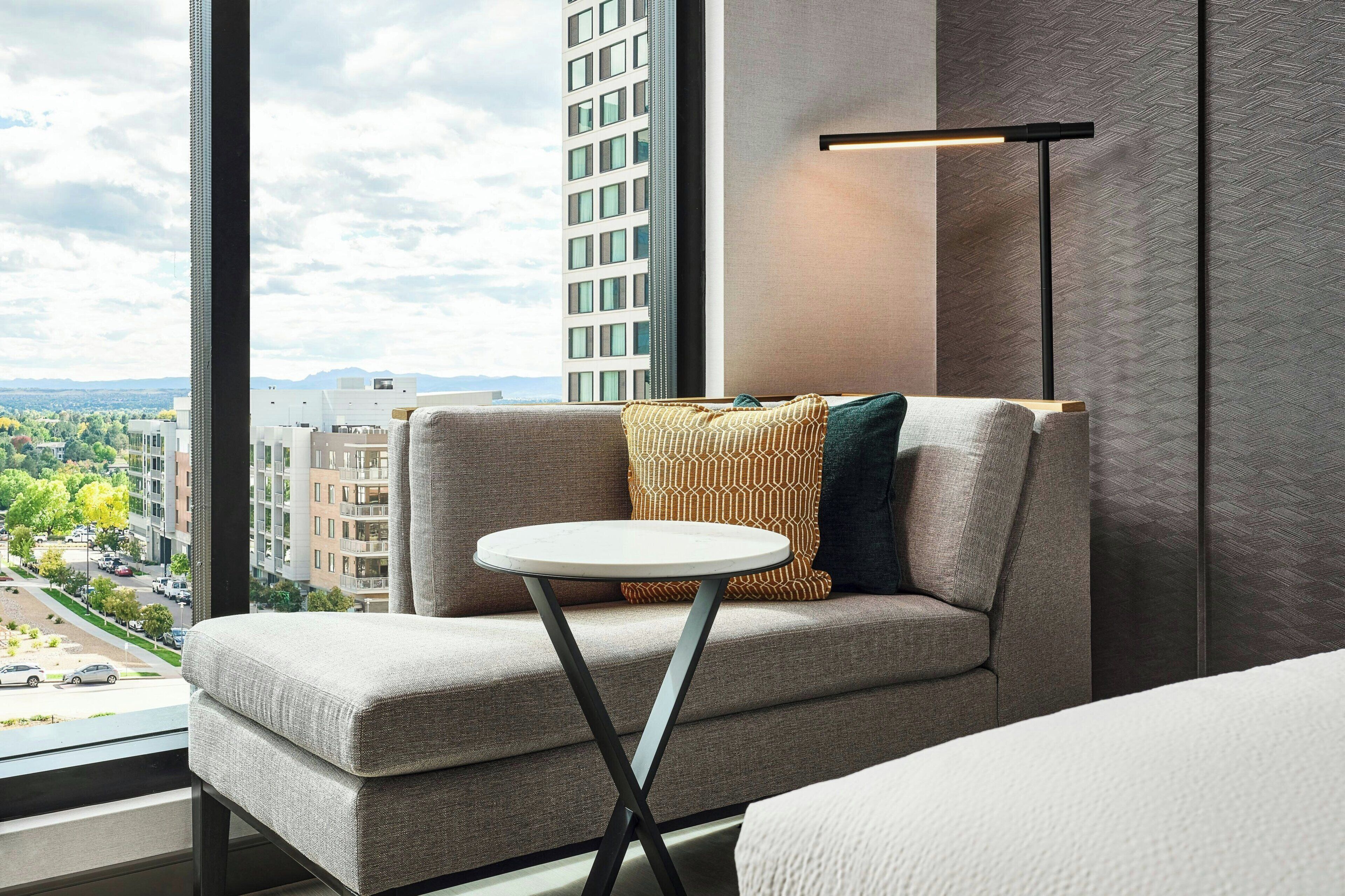 Ein modernes Hotelzimmer mit einer Couch, einem kleinen runden Tisch und Blick auf eine Stadtlandschaft durch große Fenster.