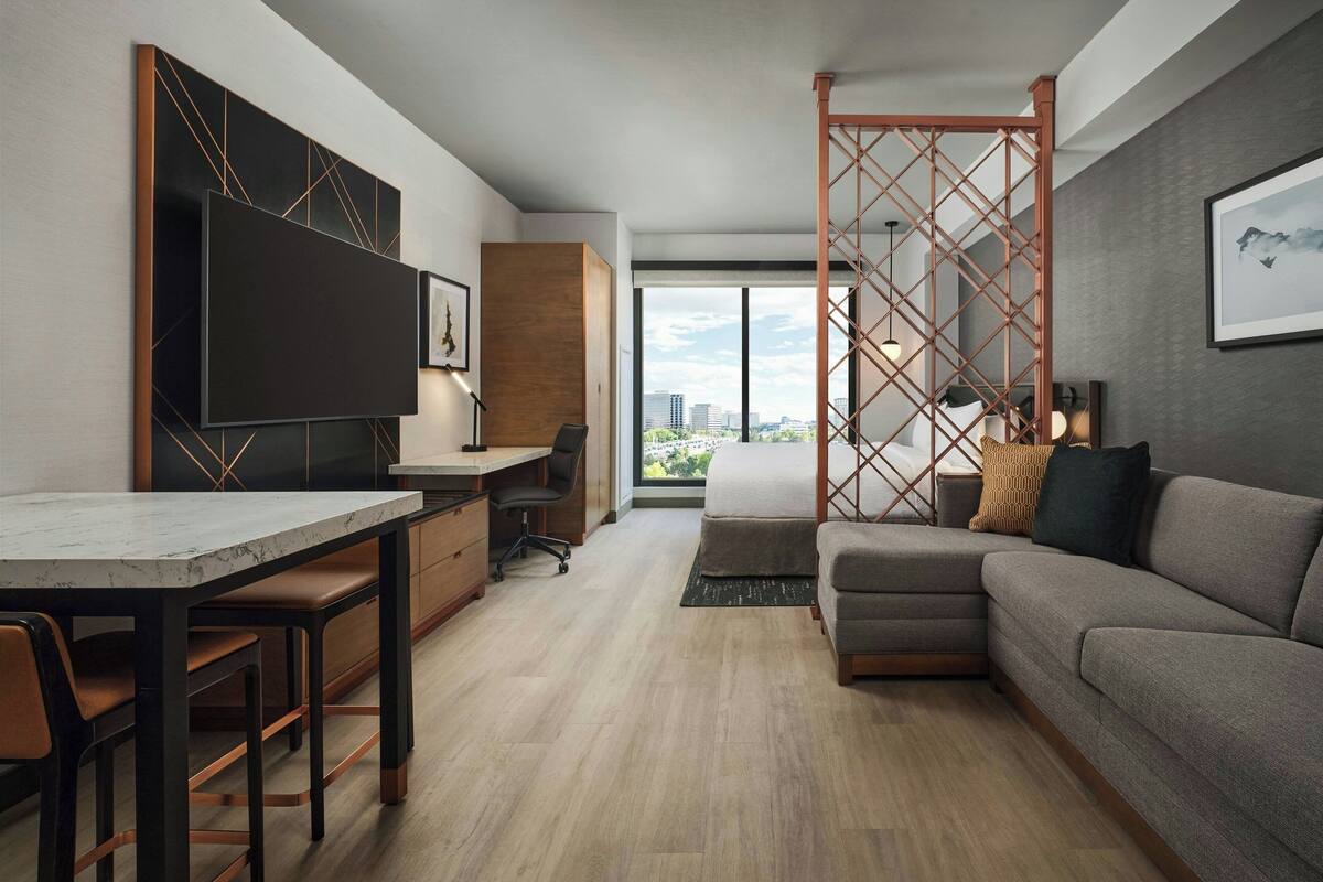 Una sala de estar moderna con un sofá, un comedor con mesa y sillas, un televisor montado en la pared y un gran ventanal con vistas a la ciudad.