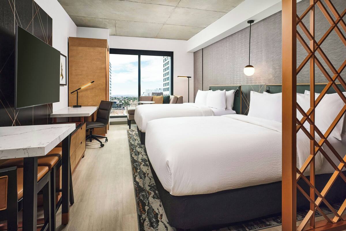 Habitación de hotel con dos camas, un escritorio con silla, televisor y ventana grande con vistas a la ciudad.