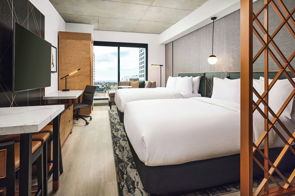 A hotel room with two beds, a desk with a chair, a TV, and a large window with a city view.