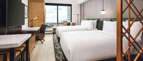 A hotel room with two beds, a desk with a chair, a TV, and a large window with a city view.