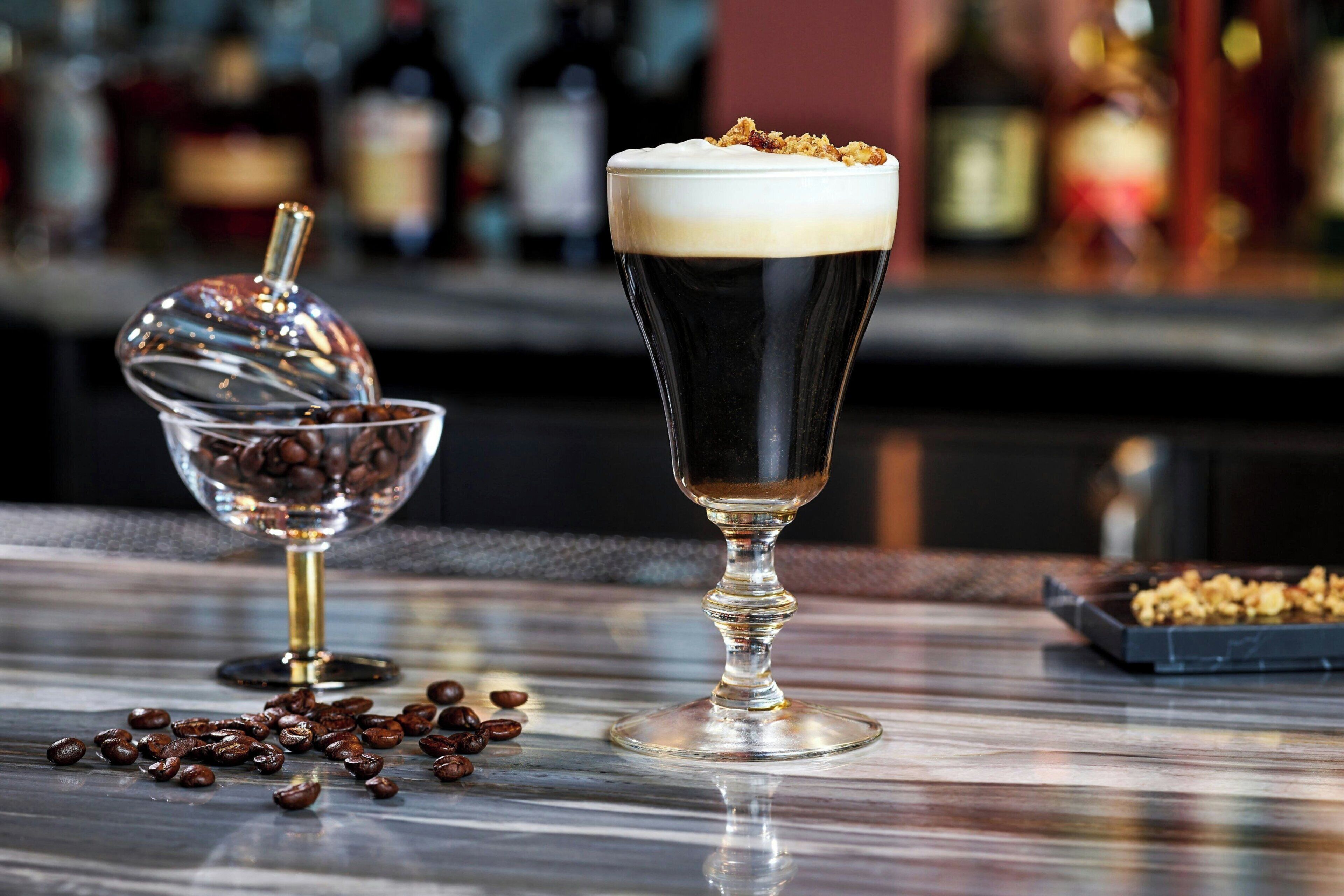 A bar setting with a glass of dark beverage, coffee beans, and a sugar bowl.