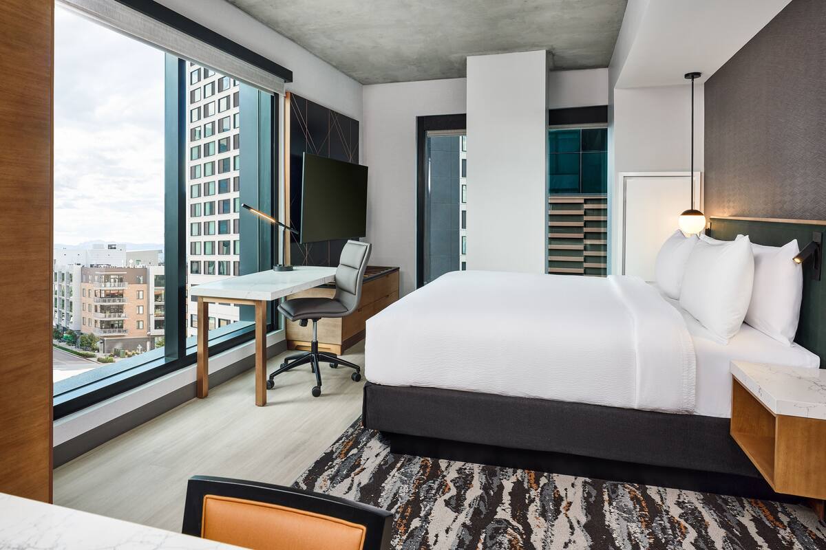 Habitación de hotel moderna con una cama grande, un escritorio y vistas a edificios a través de amplios ventanales.
