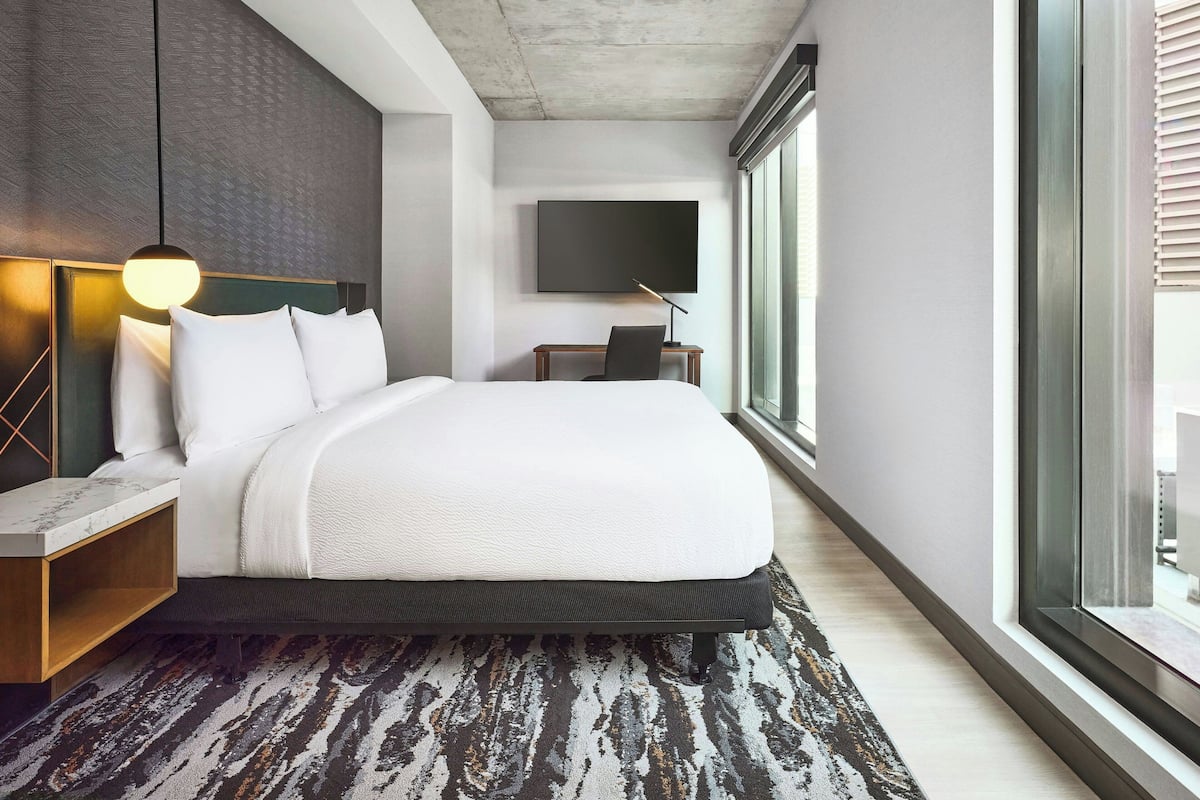 A modern hotel room with a large bed, a desk, a flat-screen TV, and a large window.