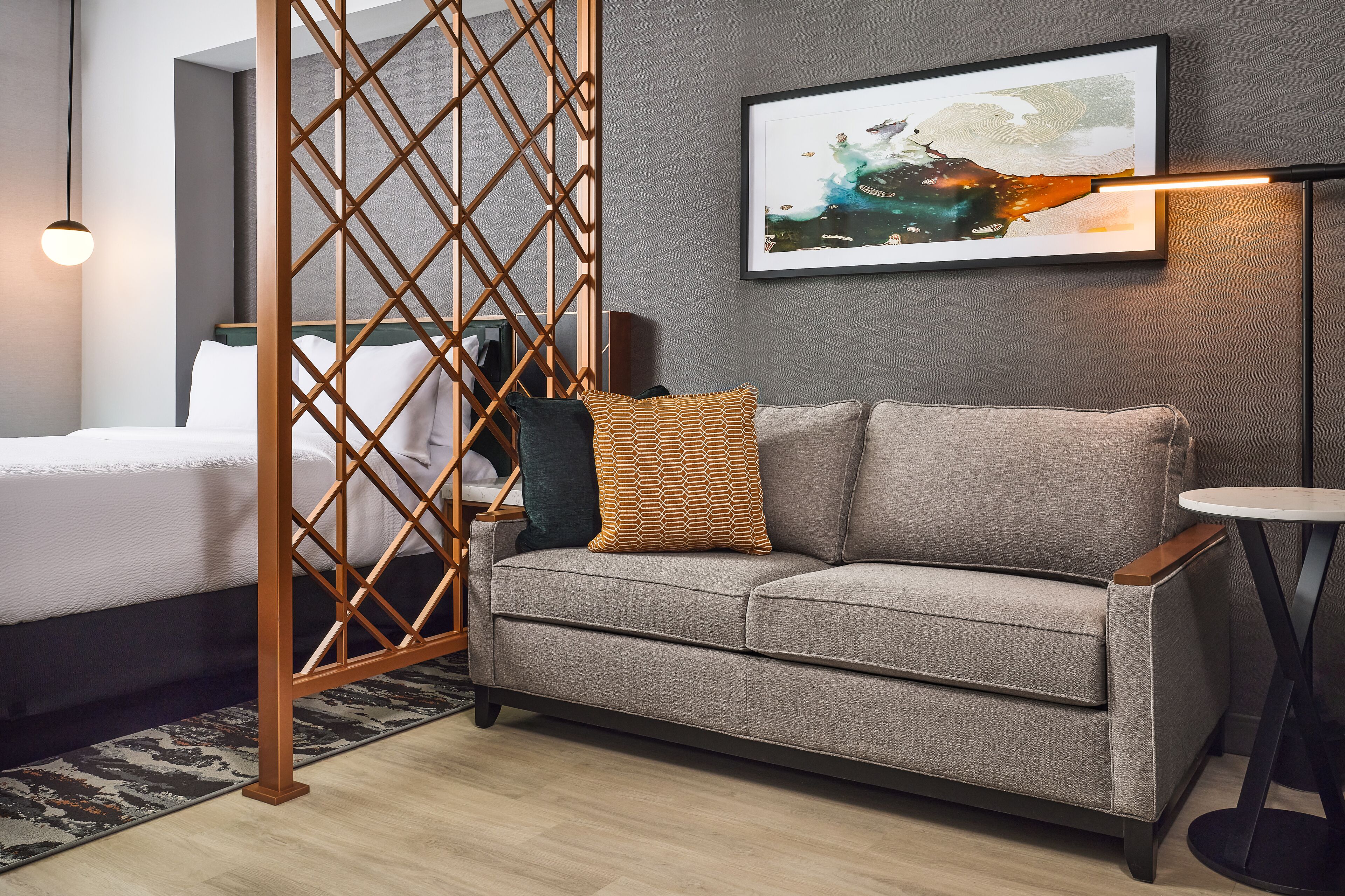 A modern hotel room with a grey sofa, a wooden partition, a framed abstract painting, and a small round table.
