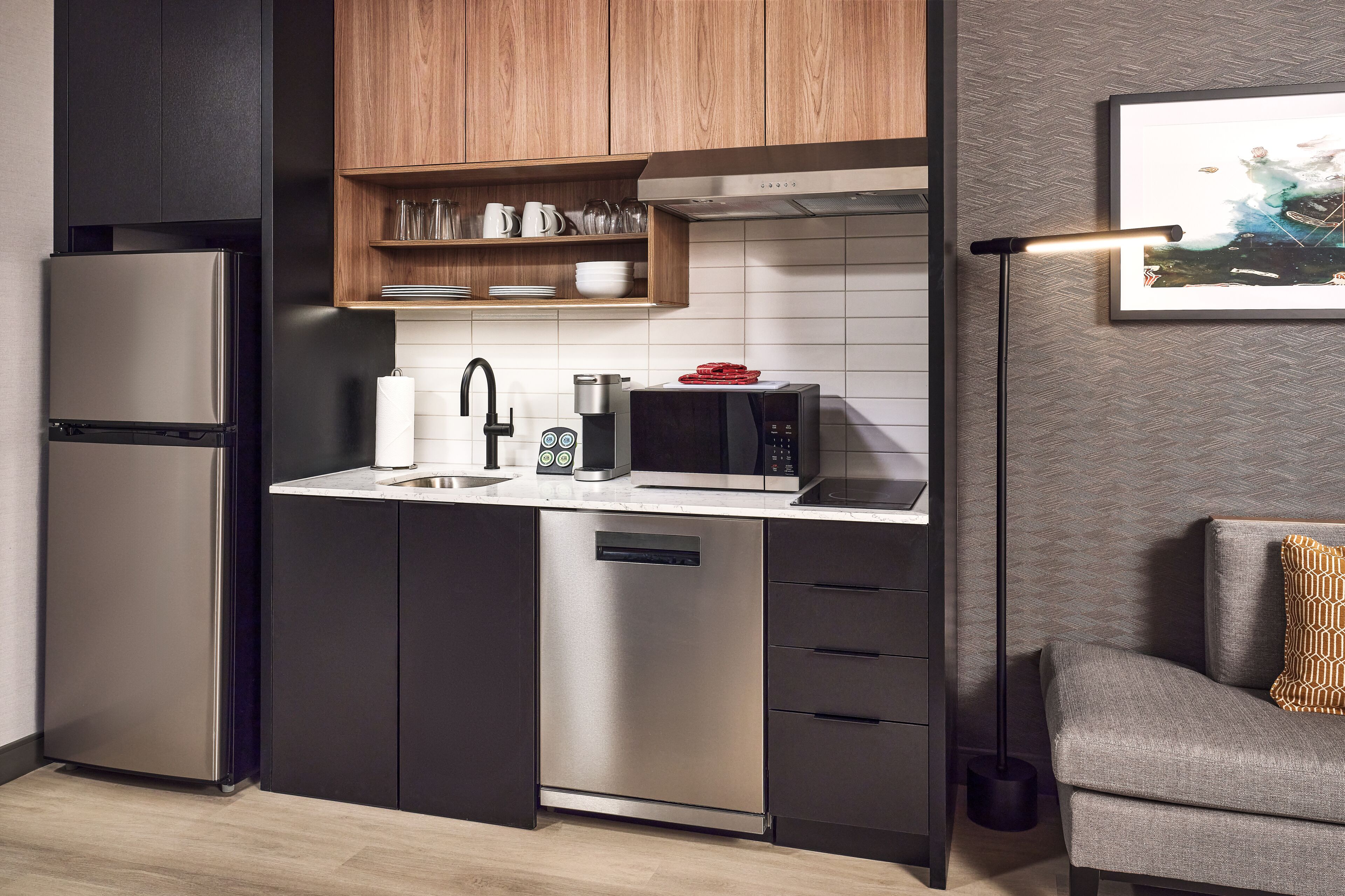 A modern kitchen with a stainless steel refrigerator, microwave, and dishwasher, wooden shelves above the sink, and a grey sofa with a patterned pillow in the corner.