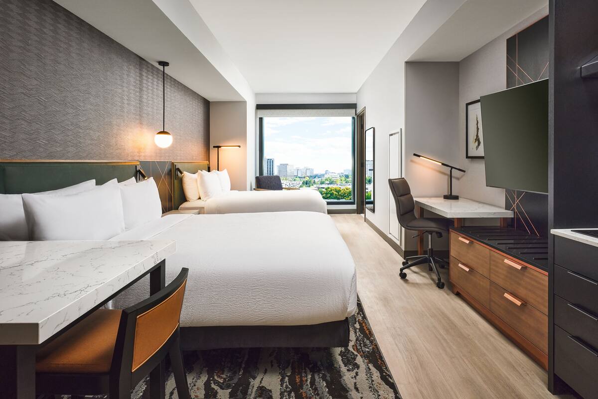 Habitación de hotel con una cama grande, un escritorio, una silla, un televisor y vista a la ciudad.