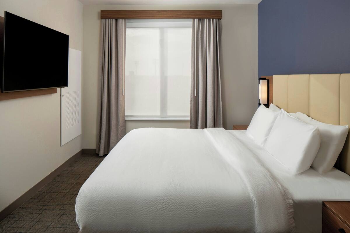 Una habitación de hotel con cama, televisor montado en la pared, ventana con cortinas y mesita de noche con lámpara.