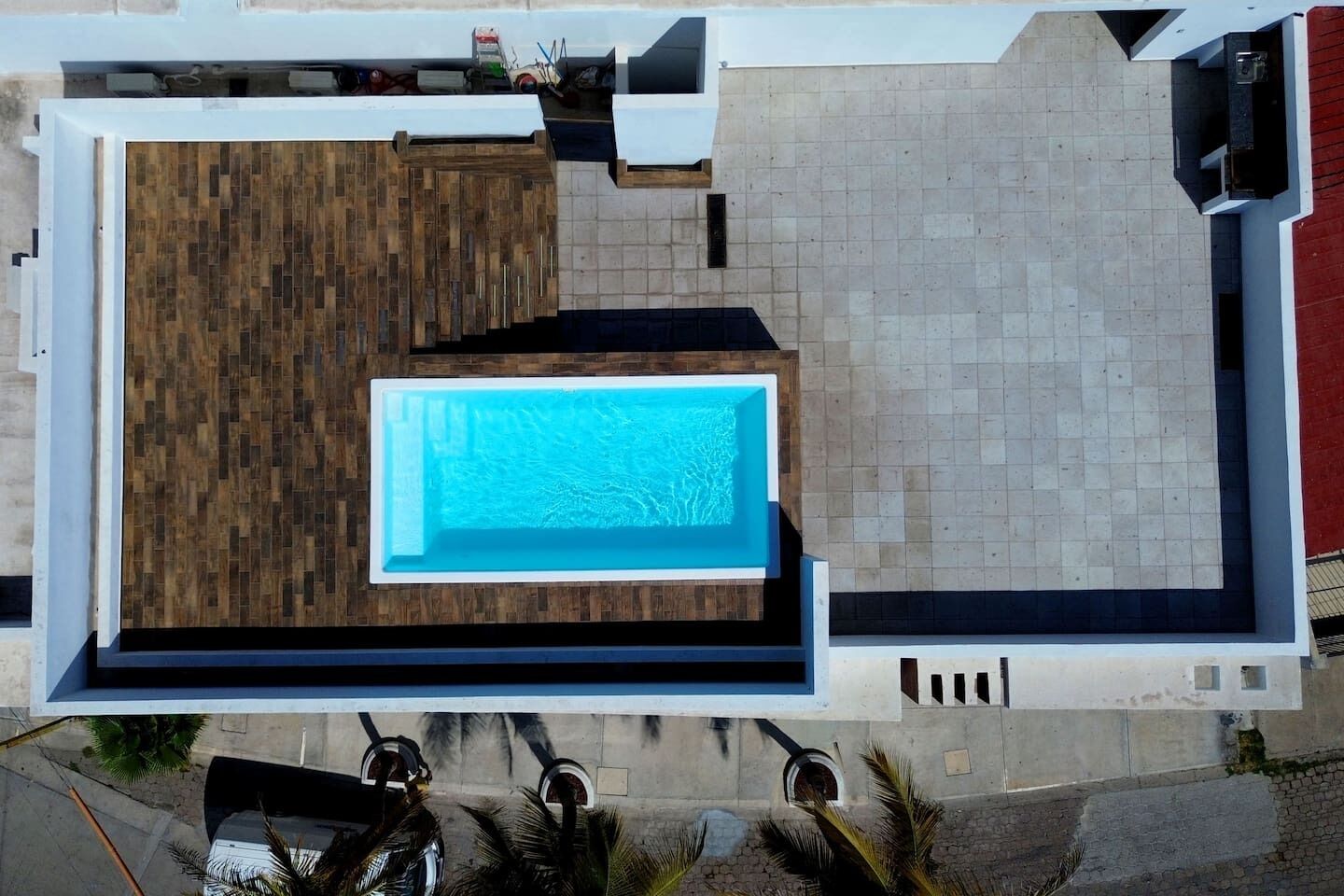 An aerial view of a modern building with a swimming pool, a wooden deck, and a tiled patio area.