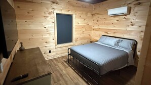 A small, cozy room with wooden walls, a single bed, a nightstand, a small table, and a window. - New Cozy Country barndominium located in Leitchfield, KY. (Leitchfield)