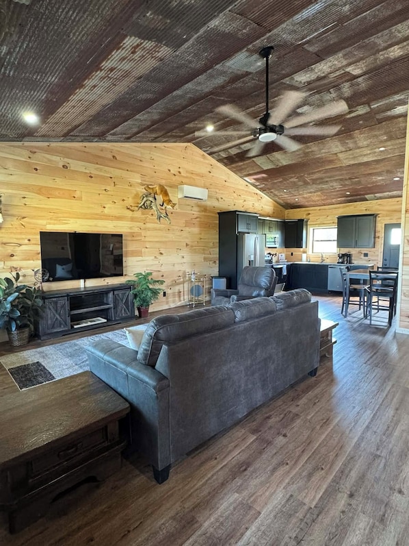 Un espacio habitable amplio con un sofá grande de color gris, techo de madera y una cocina al fondo.