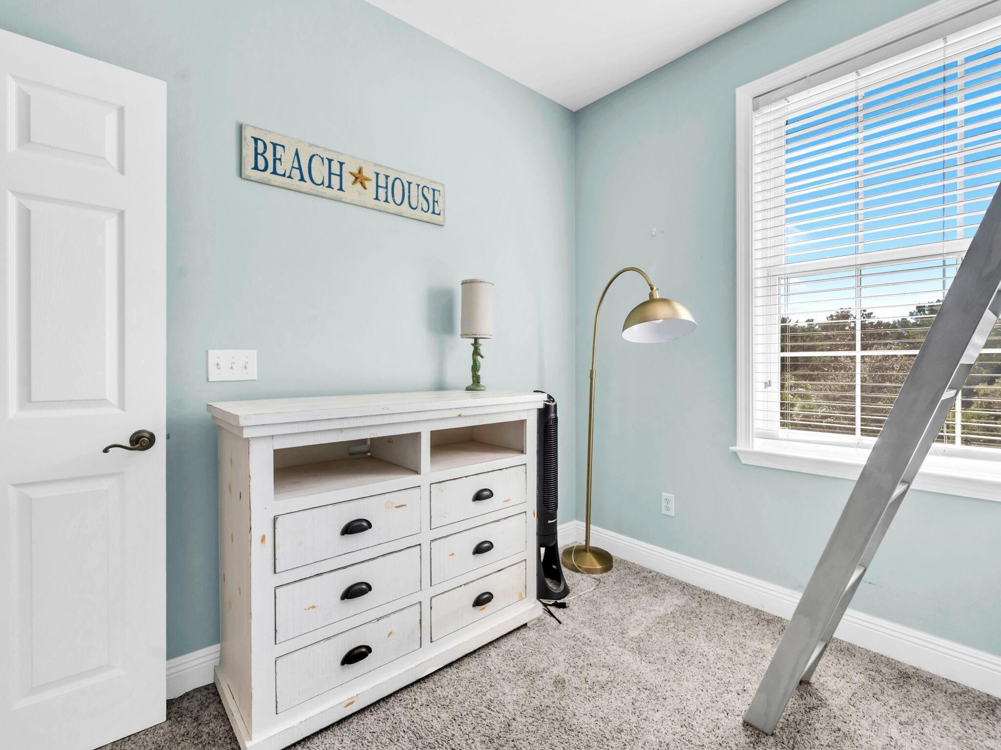Quarto com cômoda branca, luminária e uma placa com a inscrição 'BEACH HOUSE'.