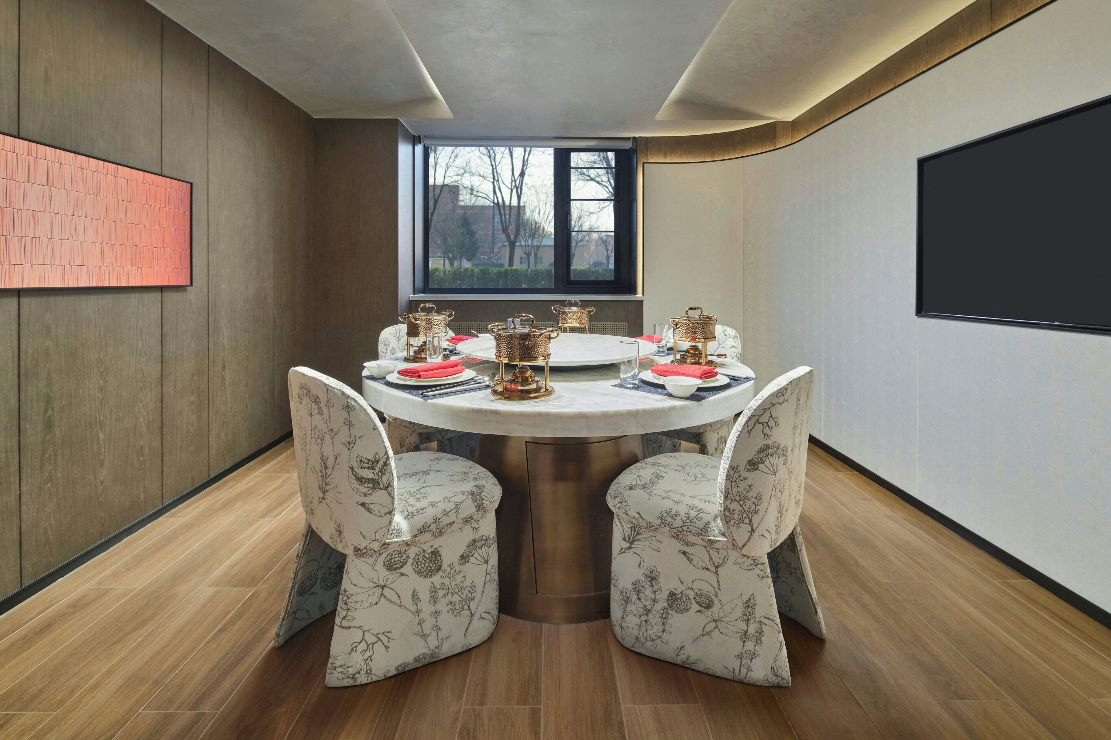 Un comedor con una mesa redonda, sillas tapizadas con motivos florales y un ventanal con vistas a árboles.
