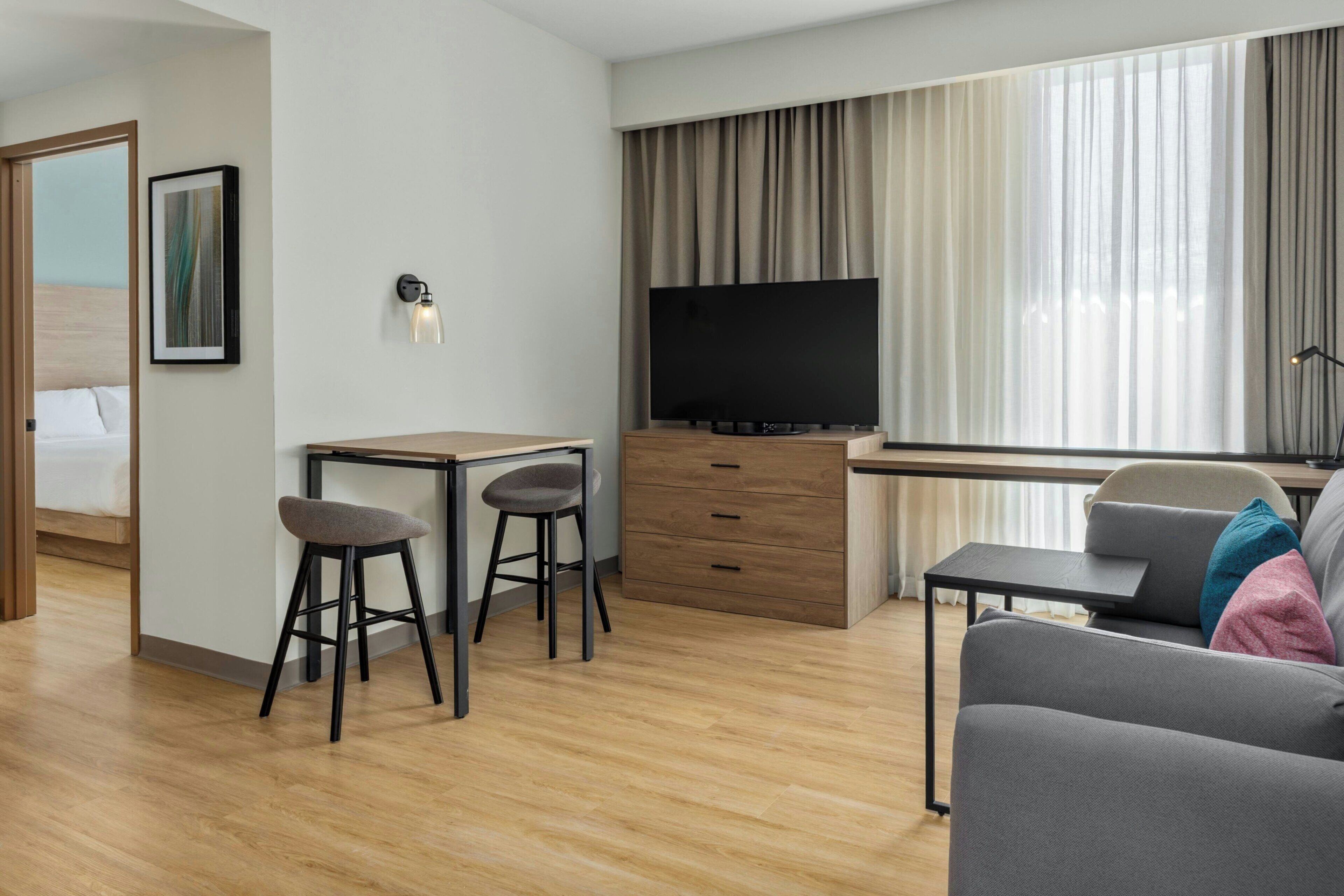 A modern hotel room with a wooden floor, a desk with two stools, a flat-screen TV, a sofa, and a bed with a wooden headboard.