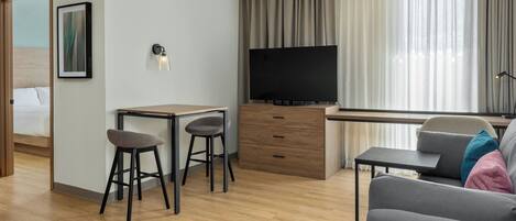A modern hotel room with a wooden floor, a desk with two stools, a flat-screen TV, a sofa, and a bed with a wooden headboard.