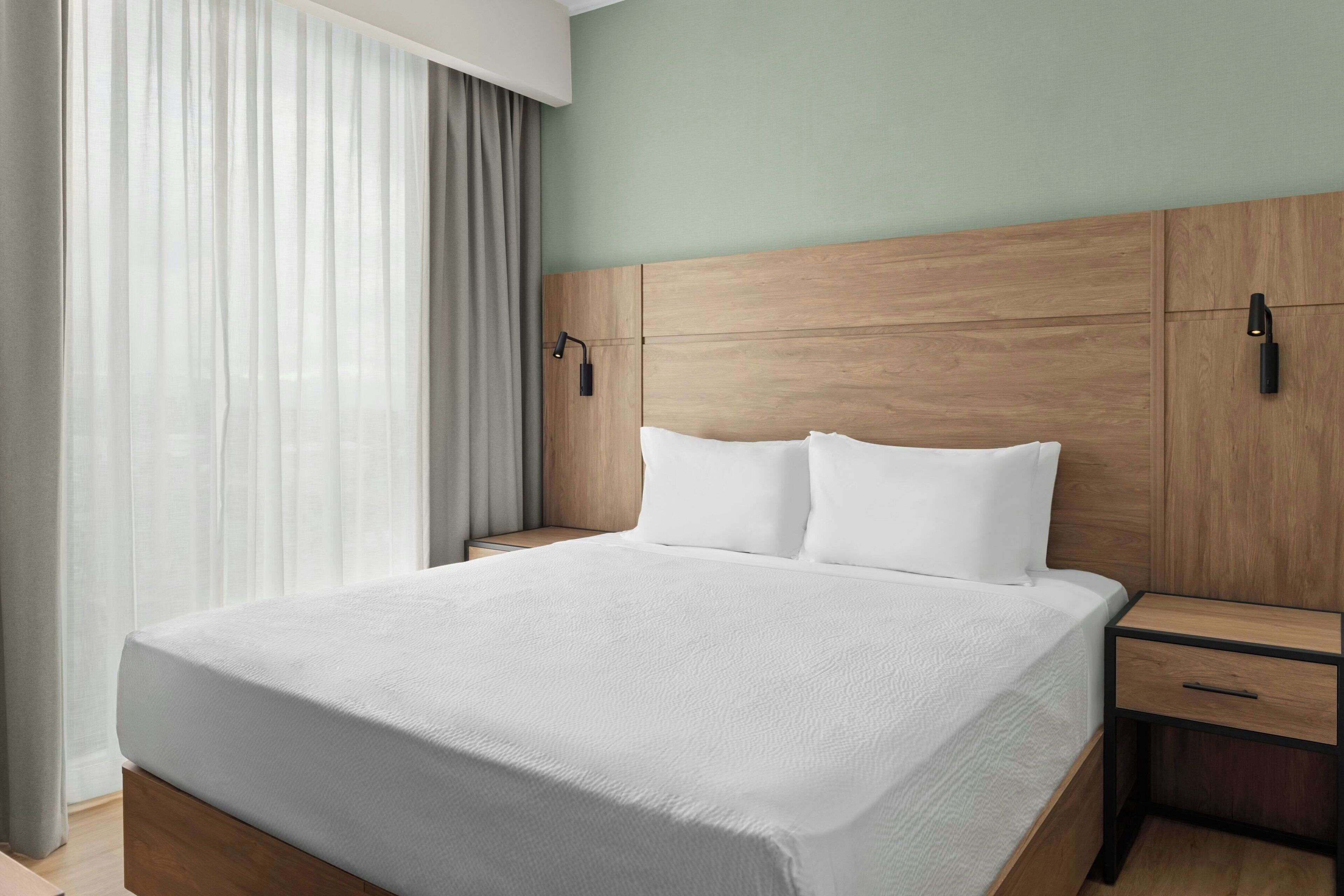 Una cama bien hecha con un cabecero de madera, una mesita de noche con cajón y una ventana con cortinas traslúcidas.