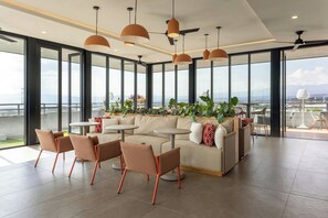 Un área de descanso moderna con sofá, sillas, mesas y lámparas colgantes. Ventanas amplias ofrecen vistas del paisaje urbano.