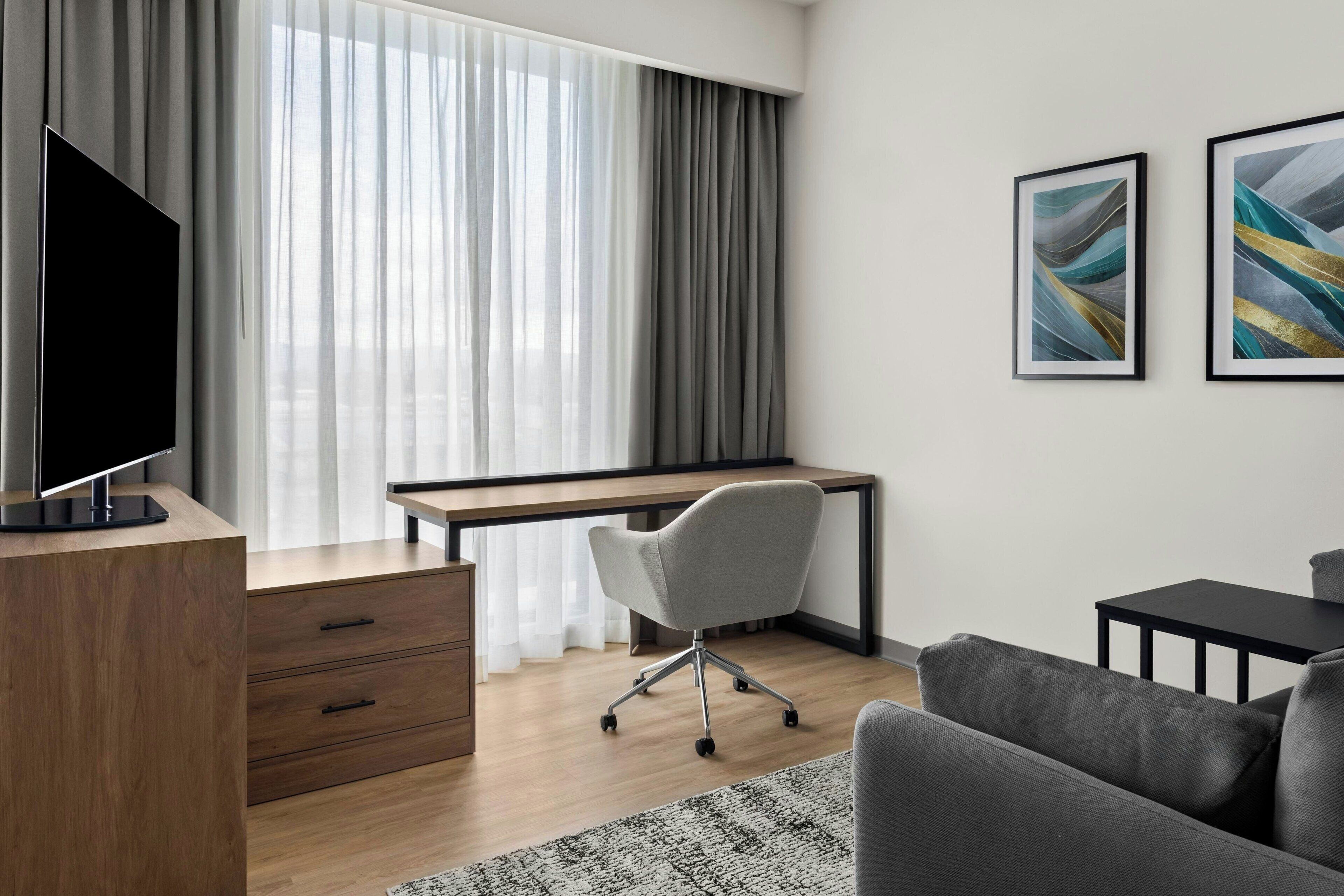 A modern hotel room with a wooden desk, a grey office chair, a flat-screen TV, a grey sofa, and two abstract paintings on the wall.