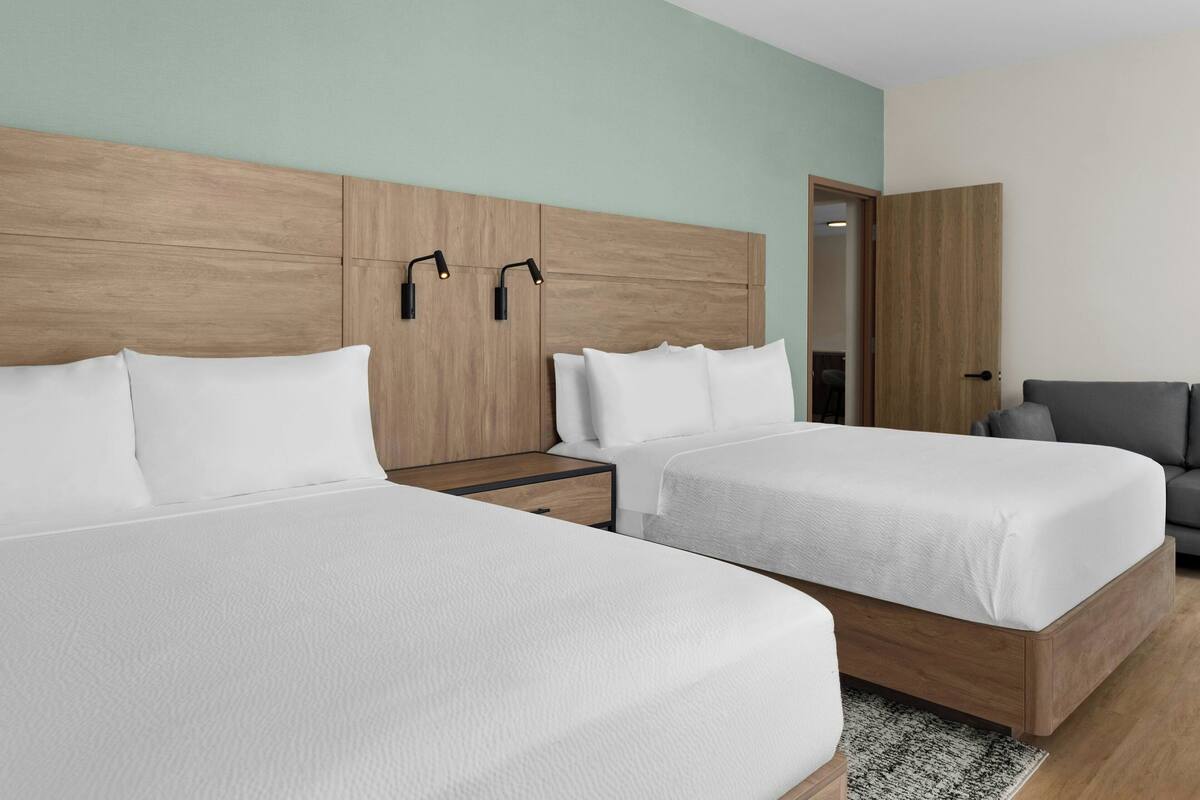 Habitación de hotel con dos camas, un cabecero de madera, un sofá gris y una puerta que da a otra habitación.